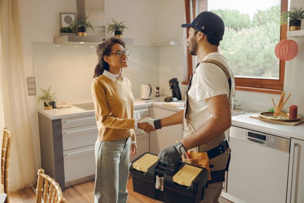 Professional repairman in uniform shaking hands with woman while standing at home kitchen Professional male handyman shaking hands with happy female client while standing at home kitchen
